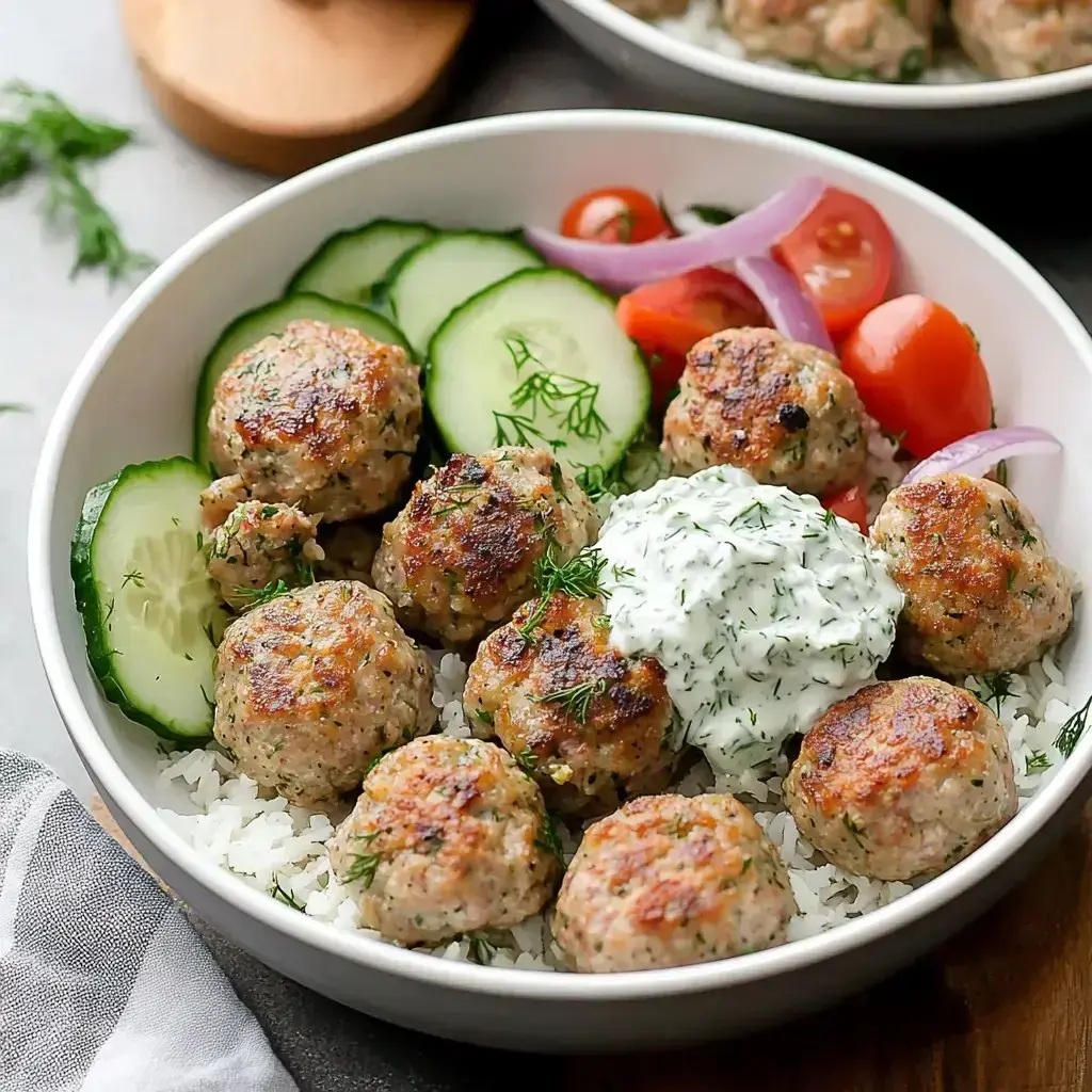 Un bol con arroz, albóndigas doradas, pepino, tomates cherry, cebolla roja y una cucharada de salsa cremosa de hierbas.