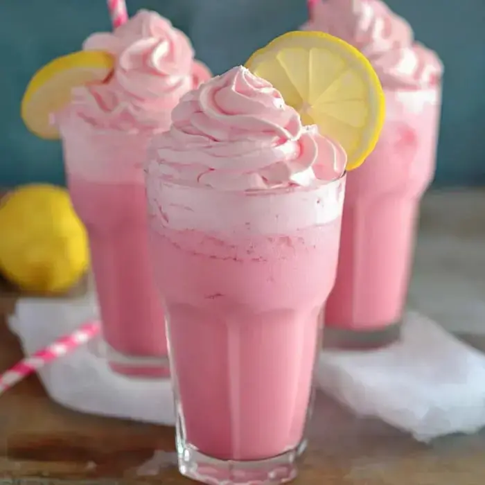 Tres vasos altos con bebidas rosadas espumosas, adornadas con crema batida y rodajas de limón; acompañados de un limón y una pajilla de papel.